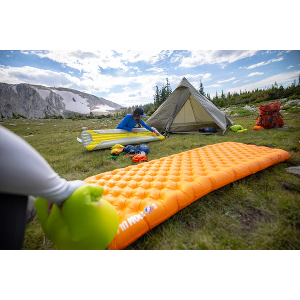 Camper setting up a sleeping pad at a backcountry campsite with a tent and mountain backdrop