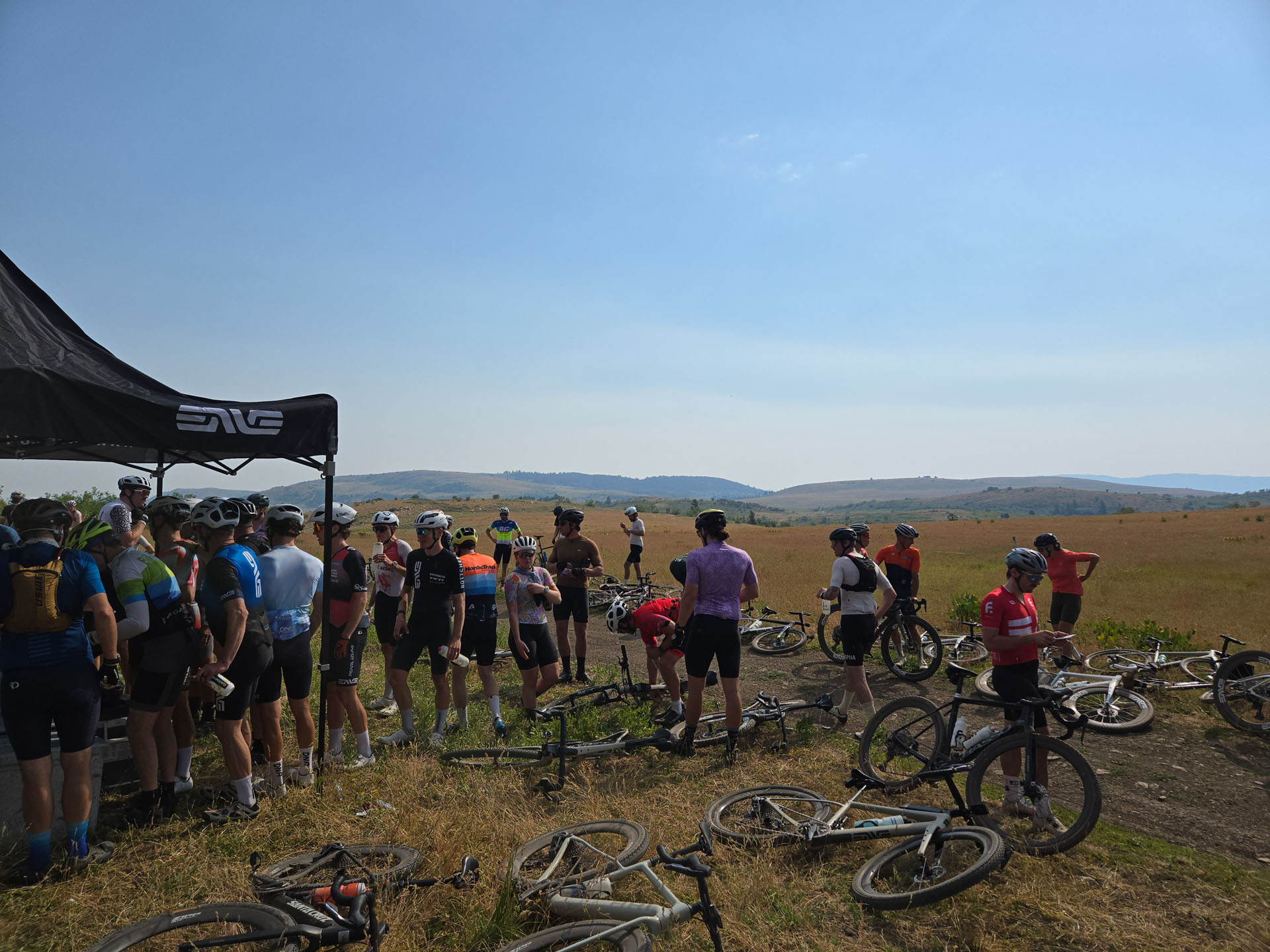 Riders regrouping at the aid-station tent, bikes down in the grass, wide vista behind